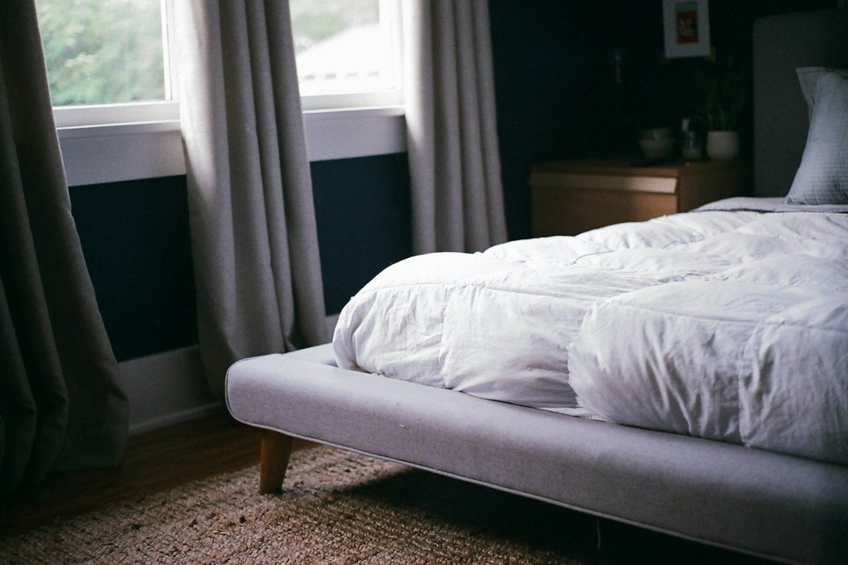 A simple bedroom with a white comforter, a low bed and soft light through the curtains